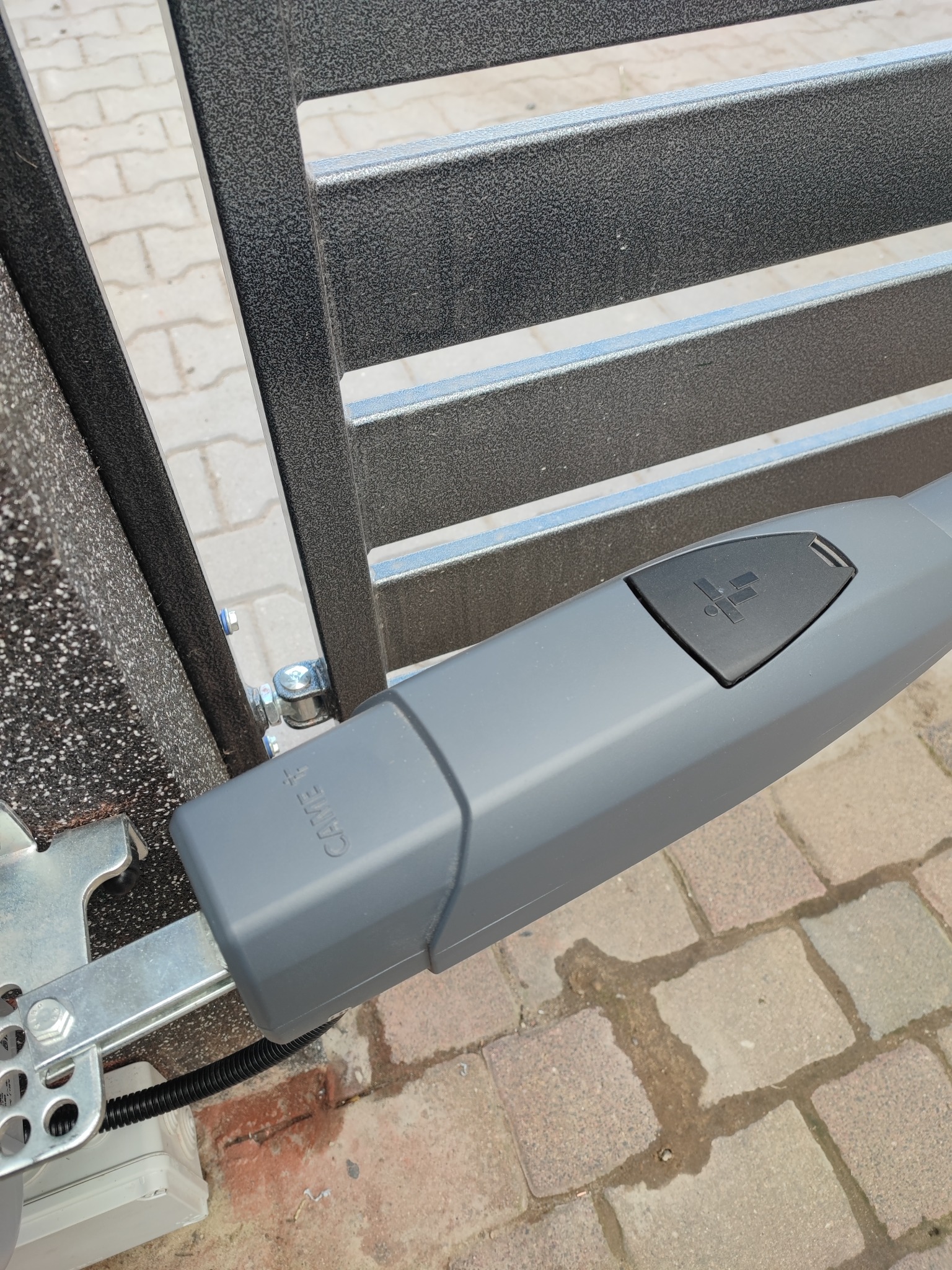 Szary siłownik bramy marki Came z widocznym logo, zamontowany do metalowej bramy posesyjnej, z okablowaniem w peszlu i kostką elektryczną, na tle kostki brukowej.