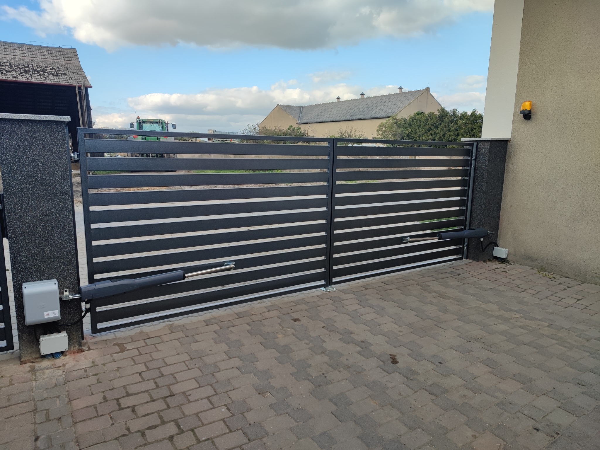 Nowoczesna brama wjazdowa z poziomymi panelami i automatyką, widoczne siłowniki, skrzynka sterownicza oraz lampa ostrzegawcza, na tle budynku gospodarczego i traktora.