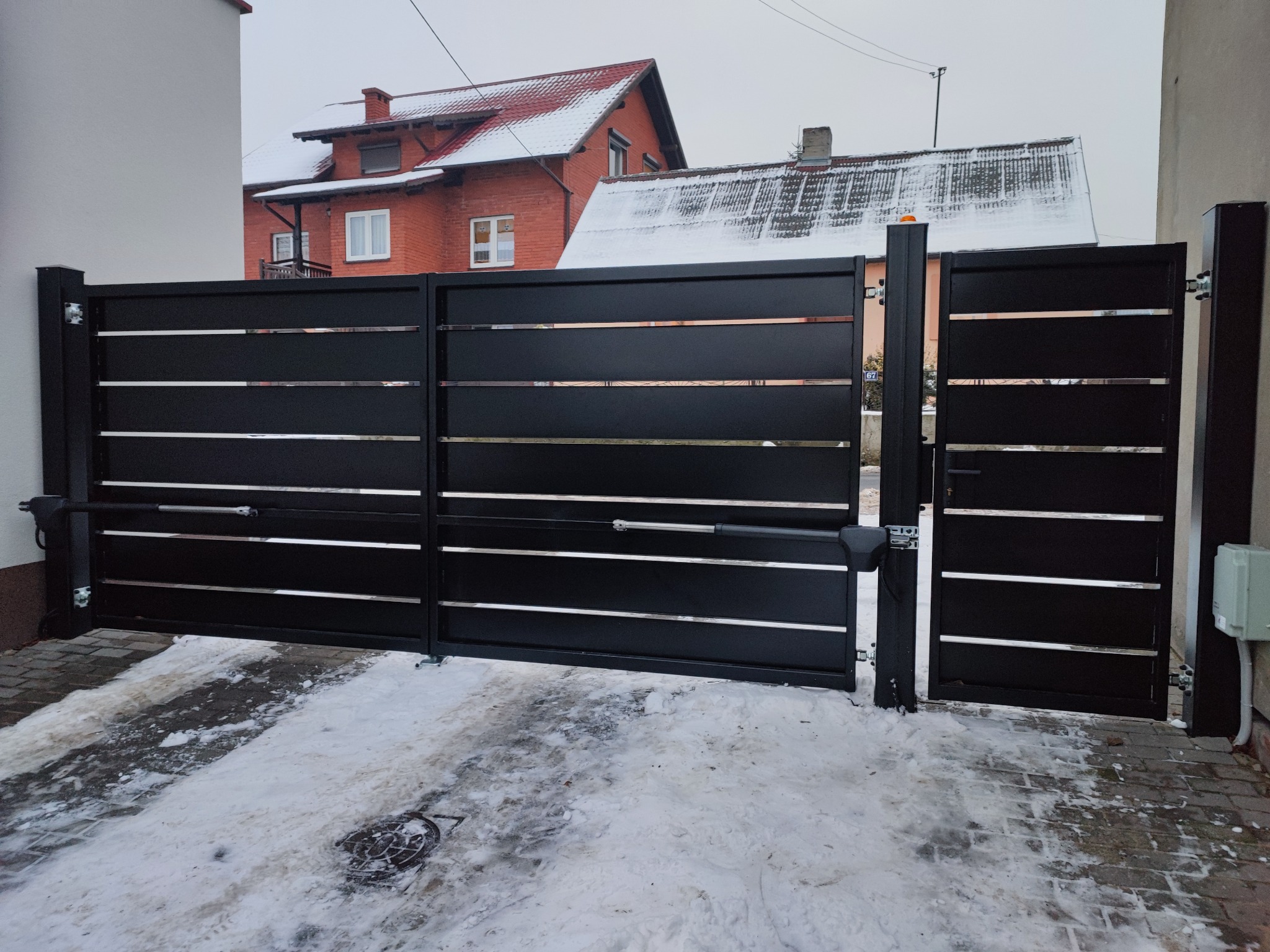 Nowoczesna, czarna brama wjazdowa z poziomymi, srebrnymi akcentami, wyposażona w siłowniki automatyczne, na tle ośnieżonego podjazdu i domu z czerwonej cegły.