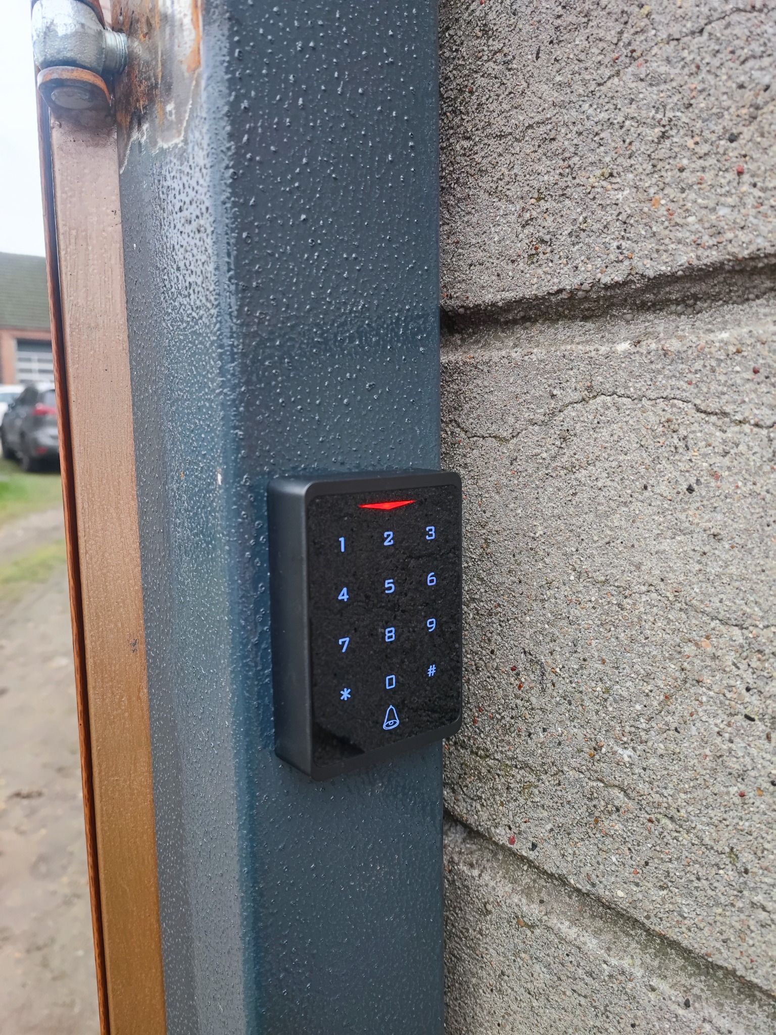 Zewnętrzny panel numeryczny z podświetlanymi przyciskami zamontowany na metalowej ramie bramy, przylegającej do betonowego słupka. Widoczne krople wody na powierzchni.