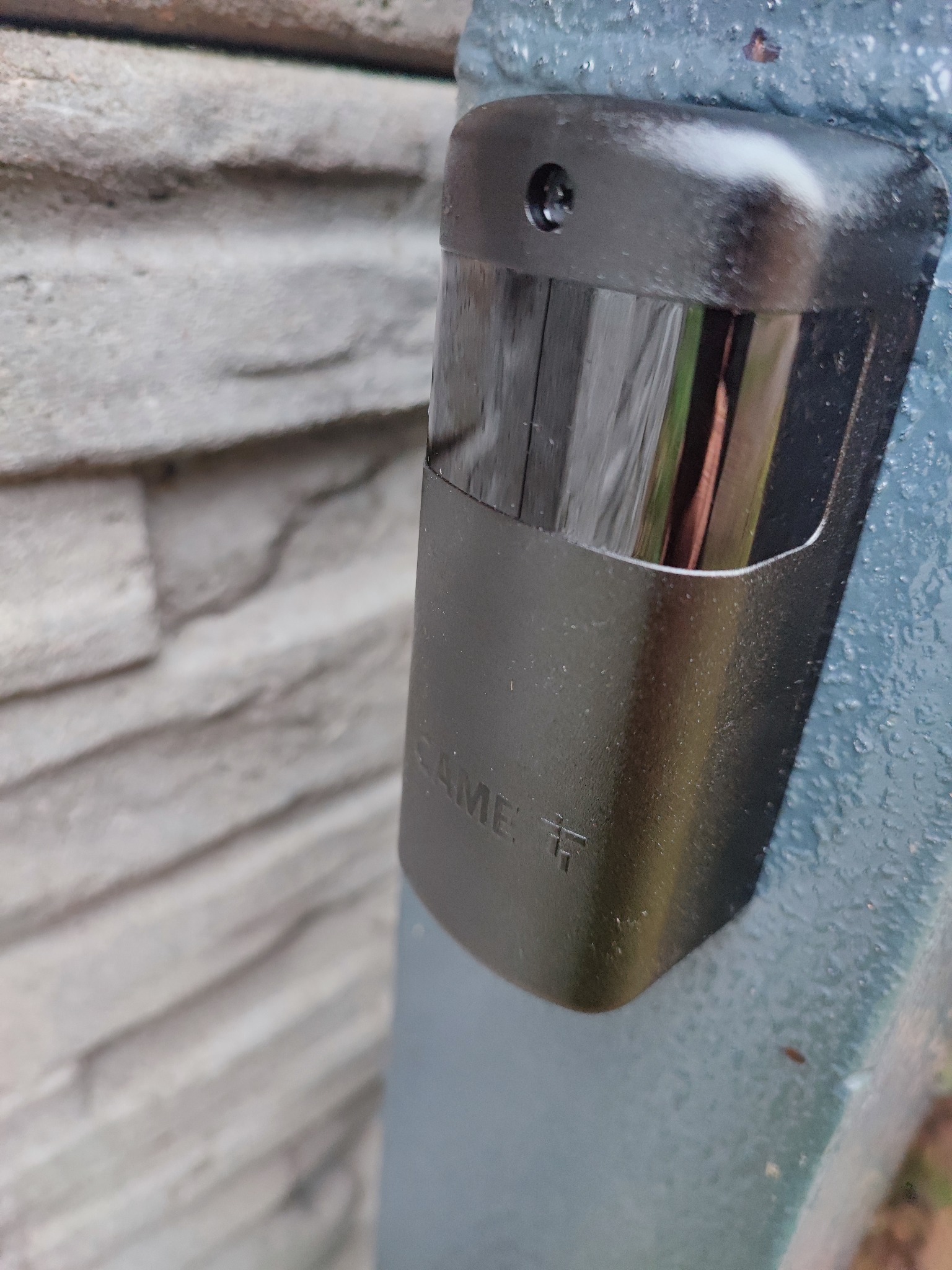 Zbliżenie na czujnik zbliżeniowy firmy CAME zamontowany na słupku bramy wjazdowej, z widocznym fragmentem murowanego ogrodzenia w tle.