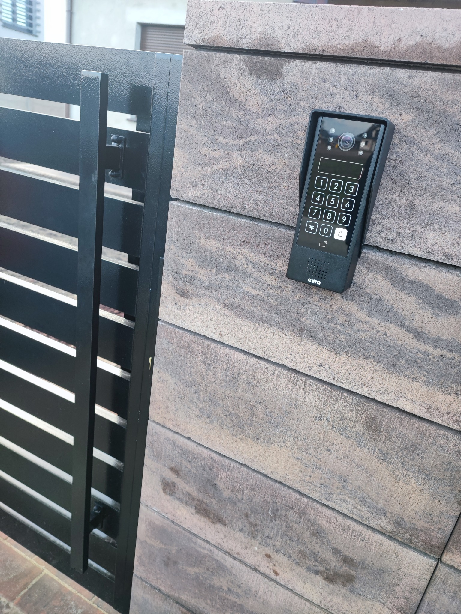 Domofon z klawiaturą numeryczną i kamerą zamontowany na murku obok metalowej bramy posesji.