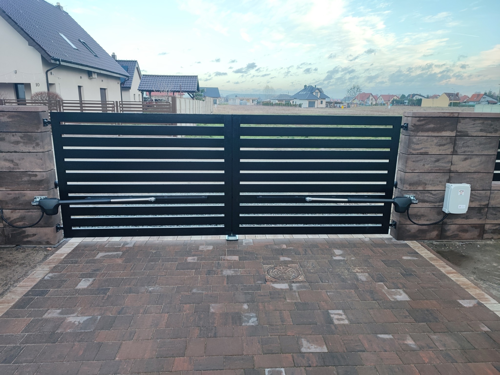 Nowoczesna, pozioma brama wjazdowa z automatyką, zamontowana na posesji z kostki brukowej, widok na domy w tle.