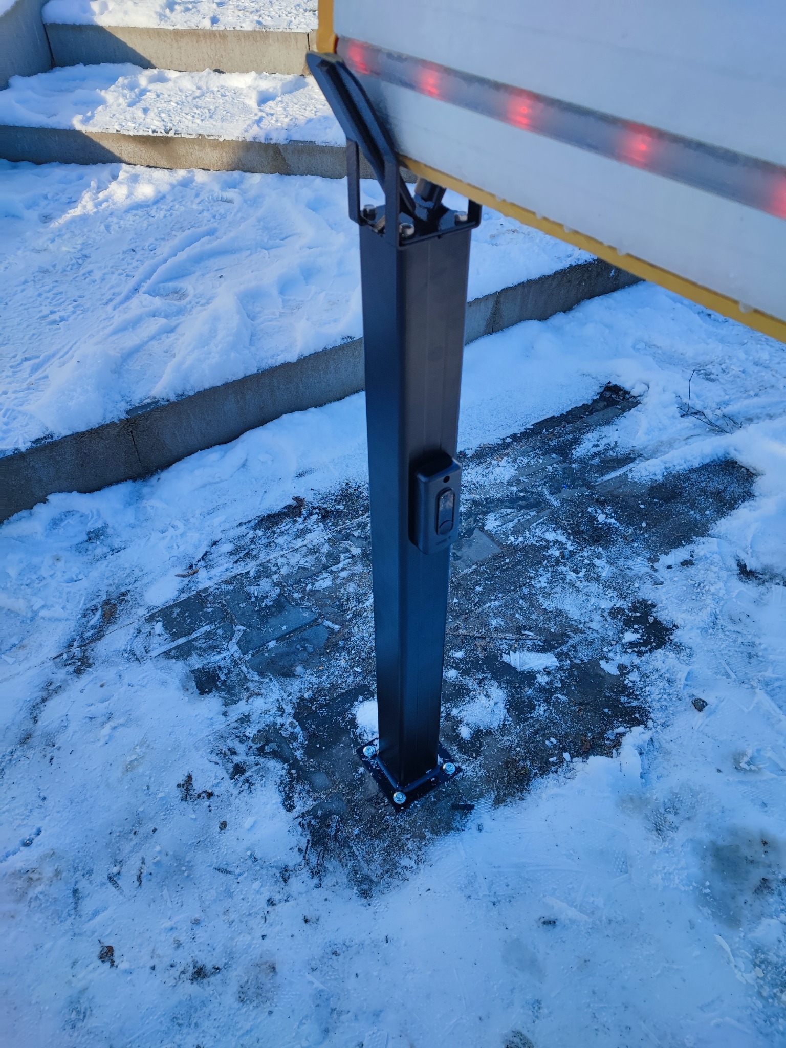 Słupek bramy automatycznej z przyciskiem, zamontowany na betonowej powierzchni pokrytej śniegiem i lodem, widoczne schody w tle.