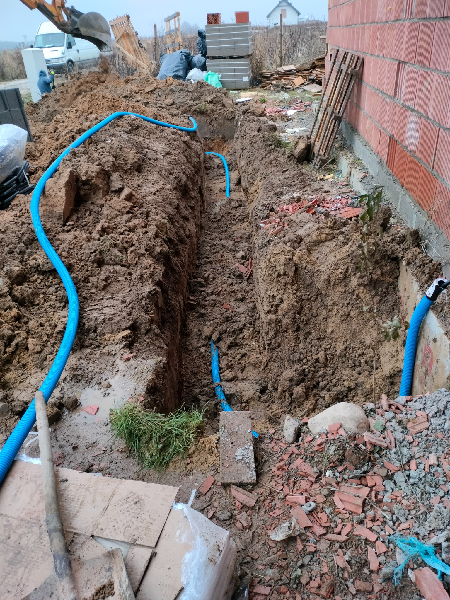 Wykopany rów w ziemi z niebieskimi rurami karbowanymi wewnątrz, widoczna ściana budynku z czerwonej cegły, koparka i materiały budowlane w tle.