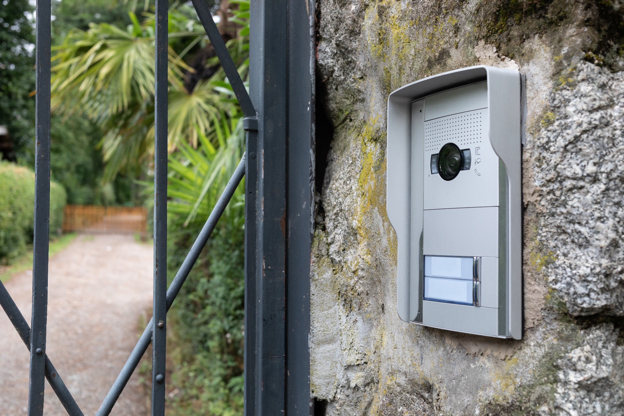 Nowoczesny wideodomofon w szarej obudowie zamontowany na kamiennym murze, widok zza metalowej bramy.