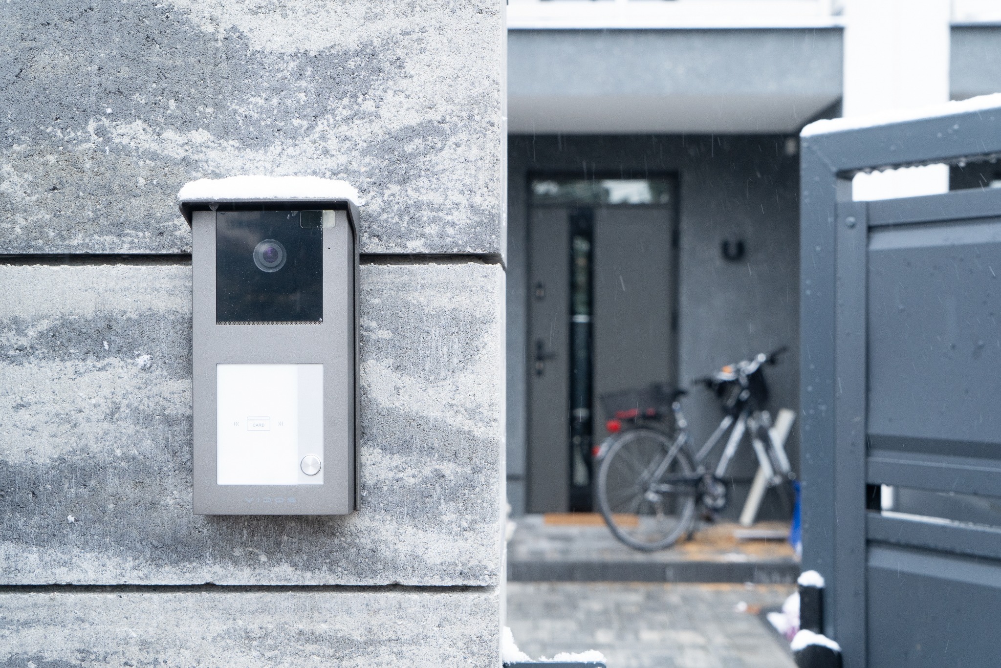 Nowoczesny wideodomofon z czytnikiem kart zbliżeniowych, zamontowany na szarej, betonowej ścianie budynku w zimowej scenerii, z widocznym rowerem i bramą w tle podczas opadów śniegu.