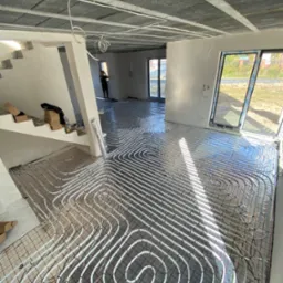 Białe rury grzewcze ułożone spiralnie na siatce w pomieszczeniu w trakcie montażu ogrzewania podłogowego, widoczne okna z widokiem na zewnątrz i osoba w oddali.