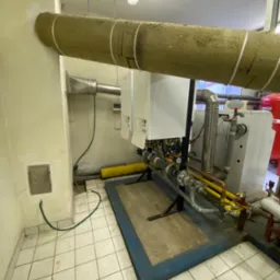 Dwa białe piece gazowe zamontowane na ścianie w kotłowni, widoczne rury, izolacja na rurze, manometry i inna armatura. Podłoga wyłożona białymi płytkami.