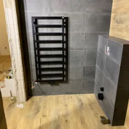 Czarny, drabinkowy grzejnik łazienkowy zamontowany na szarej, kwadratowej płytce ściennej, widoczny w trakcie montażu instalacji w łazience z drewnopodobną podłogą i zabudową podtynkową WC...