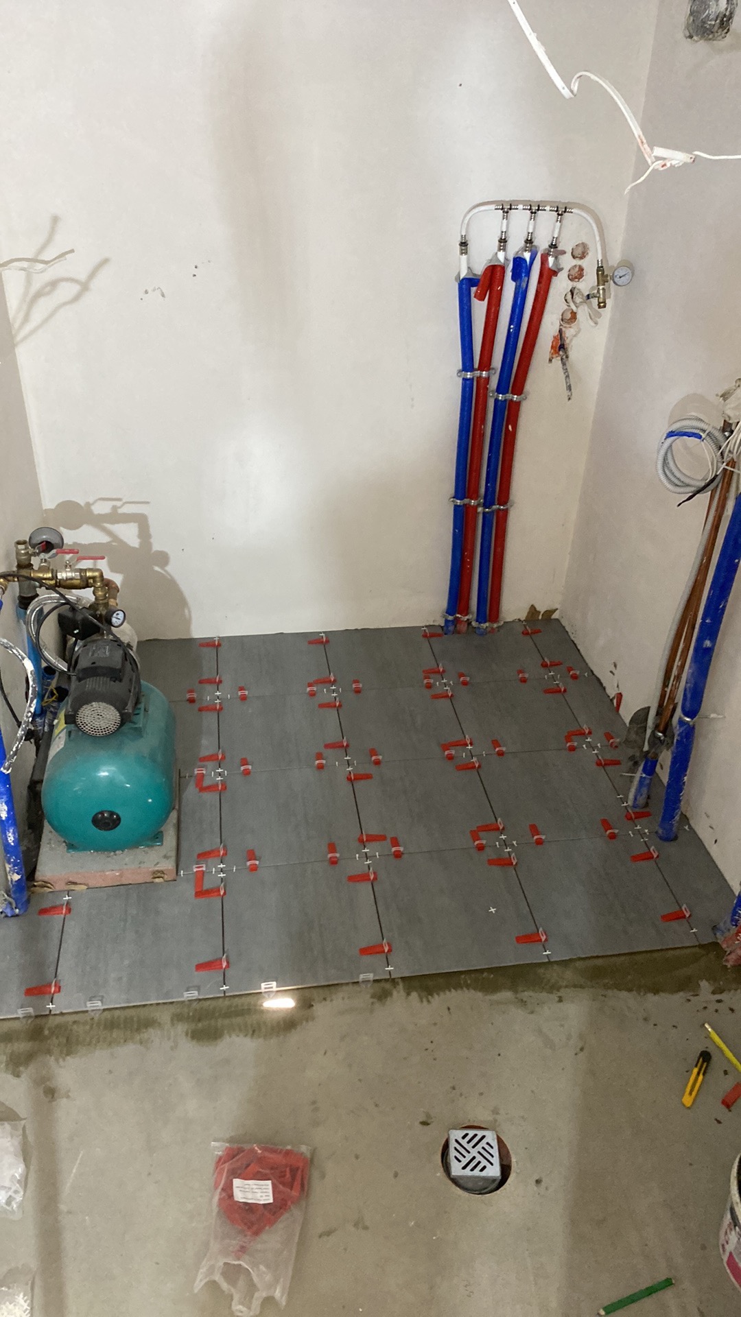 Montaż szarych płytek podłogowych z użyciem czerwonych klipsów poziomujących, widoczne rury instalacji wodnej w kolorach niebieskim i czerwonym oraz hydrofor, surowe ściany w tle.