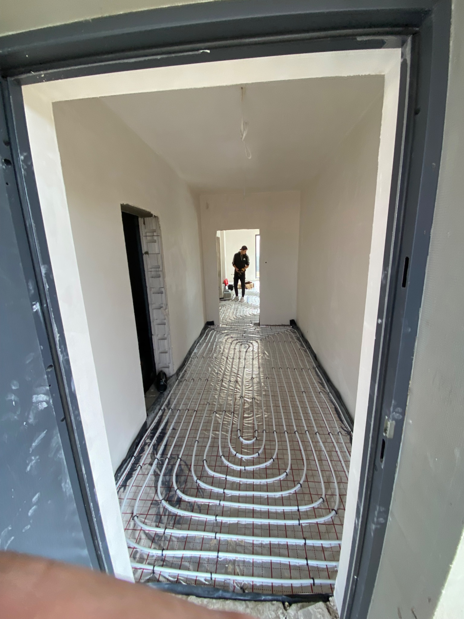 Korytarz w trakcie instalacji ogrzewania podłogowego, widoczne białe rurki ułożone na folii izolacyjnej z czerwoną siatką, w tle pracownik w czarnym ubraniu.