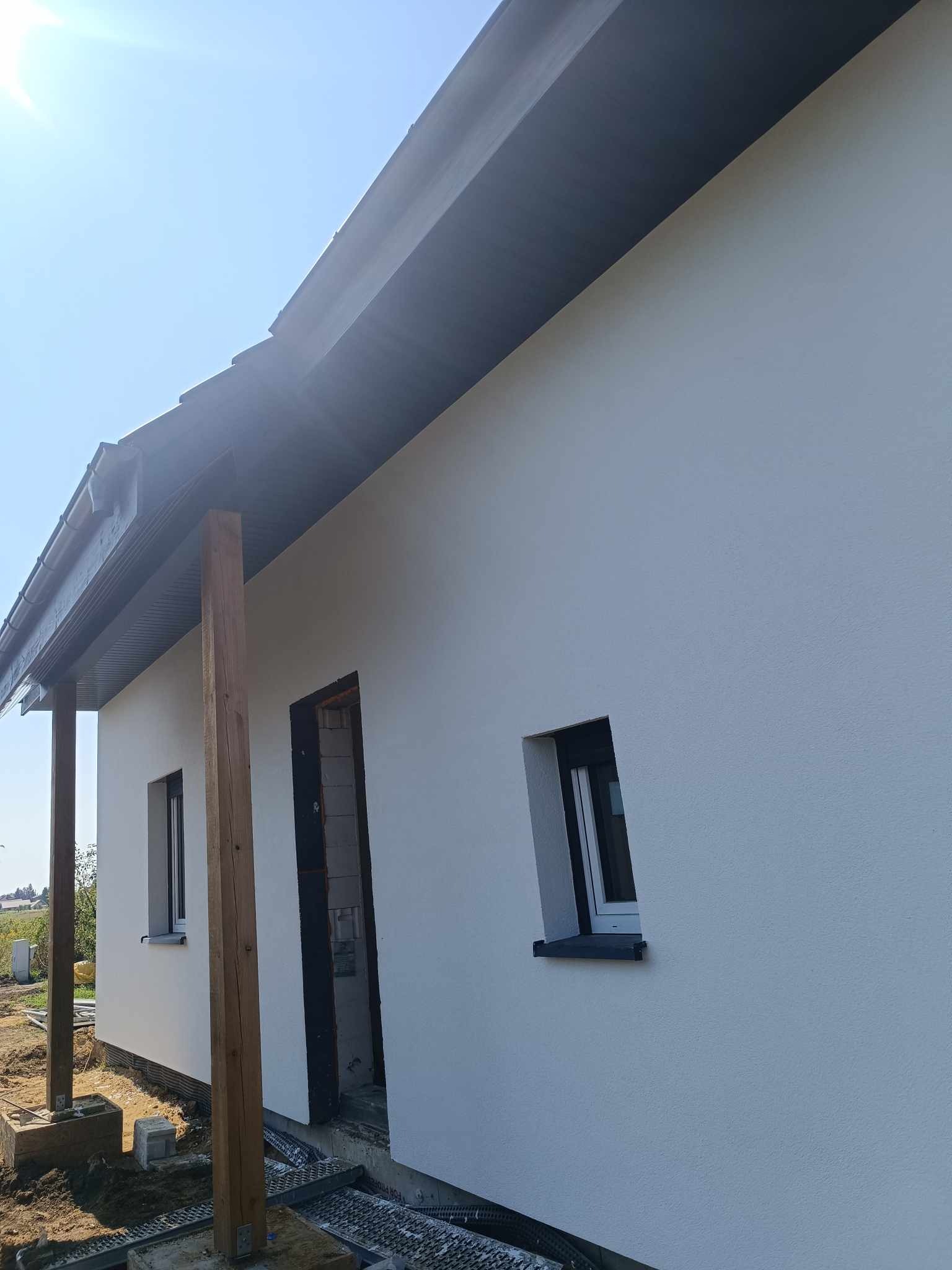 Świeżo otynkowana, biała elewacja domu jednorodzinnego z ciemnymi oknami i drewnianymi słupami wspierającymi zadaszenie, widoczne detale wykończeniowe i przygotowania do dalszych prac.
