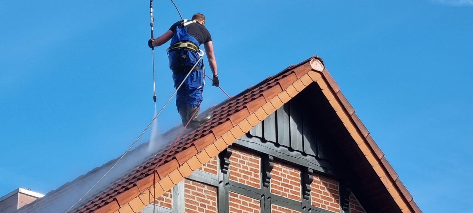 Pracownik w kombinezonie czyści dach z czerwonej dachówki za pomocą myjki ciśnieniowej. Widoczny fragment ściany budynku z cegły i drewnianych elementów.