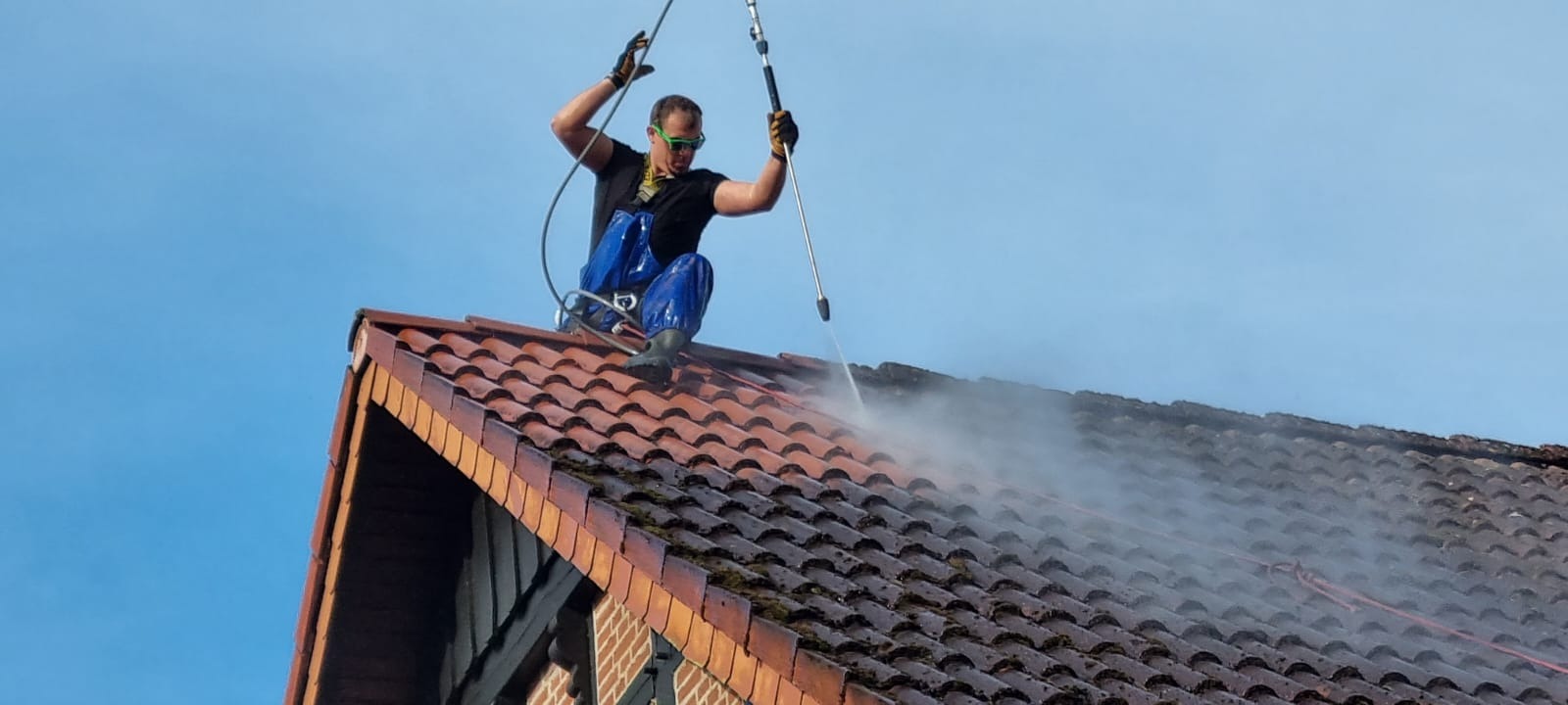 Pracownik w kombinezonie, z zabezpieczeniem, czyści dachówkę z mchu za pomocą myjki ciśnieniowej. Widoczny strumień wody i czyszczona powierzchnia dachu.