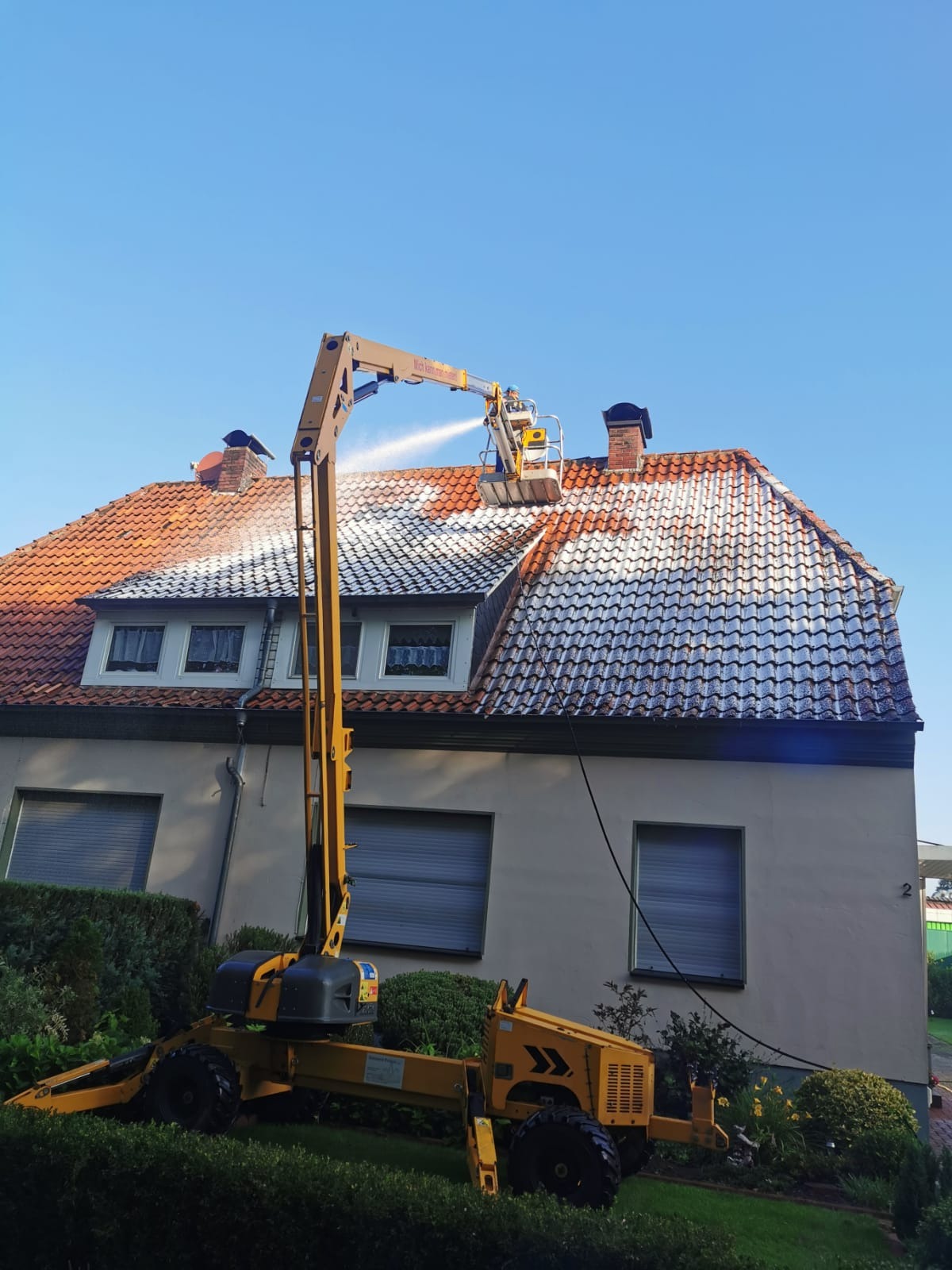 Częściowo umyty dach z czerwonej dachówki, czyszczony z podnośnika koszowego. W tle jasna elewacja domu i błękitne niebo.