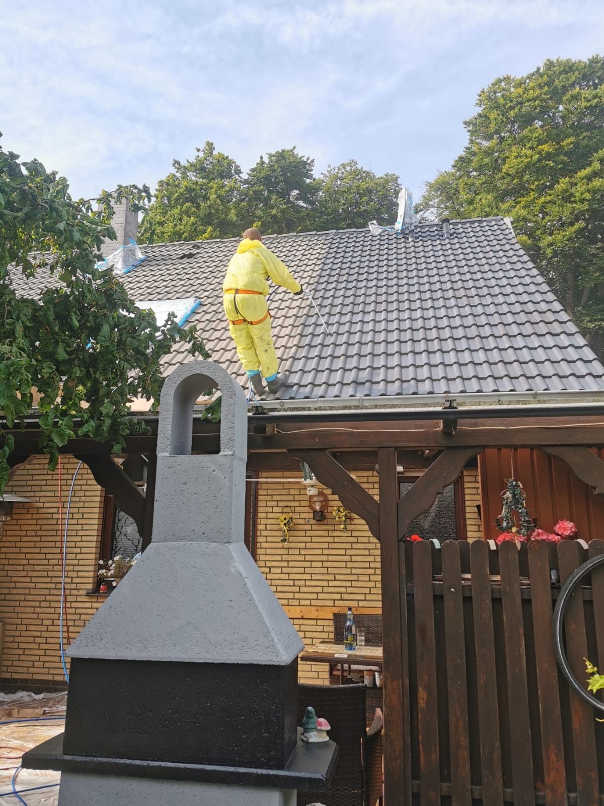 Pracownik w żółtym kombinezonie czyści dach z szarej dachówki za pomocą lancy ciśnieniowej. Widoczny fragment muru z cegły i drewniana konstrukcja altany.