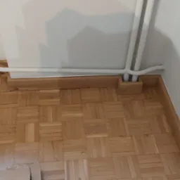 Parkiet w jodełkę z drewnianą listwą przypodłogową, biały grzejnik żeberkowy i rury grzewcze przy białej ścianie.