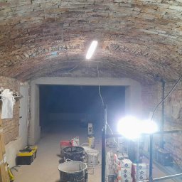 ROBERT MYKYTA RMD - Wnętrze piwnicy z ceglanym sklepieniem łukowym w trakcie prac remontowych, widoczne wiadra, zaprawy, narzędzia i oświetlenie budowlane.