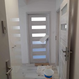 ROBERT MYKYTA RMD - Świeżo pomalowane wnętrze z dwiema parami drzwi w odcieniach bieli i szarości, wiadro z farbą na podłodze, zabezpieczone drzwi folią malarską.