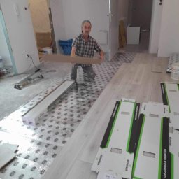 ROBERT MYKYTA RMD - Układanie paneli podłogowych: mężczyzna w koszuli w kratę przygotowuje panel, widoczne rozłożone podkłady i kartony po panelach marki Wenninger, częściowo ułożona podłoga z paneli i fragment...