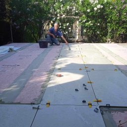 ROBERT MYKYTA RMD - Układanie jasnoszarych płytek na zewnątrz, z użyciem poziomujących klipsów i klinów, na tle kwitnących bzów i domu z czerwoną dachówką.