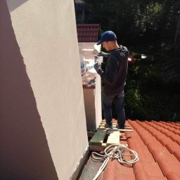 ROBERT MYKYTA RMD - Pracownik w niebieskiej czapce nakłada szpachelką jasną masę na komin z zewnątrz, stojąc na prowizorycznym rusztowaniu z desek na czerwonym dachu.