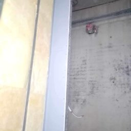 ROBERT MYKYTA RMD - Ściana w trakcie wykańczania: izolacja z wełny mineralnej, płyta gipsowo-kartonowa na obrzeżu otworu drzwiowego, instalacje elektryczne w peszlu widoczne na szarej, surowej ścianie.