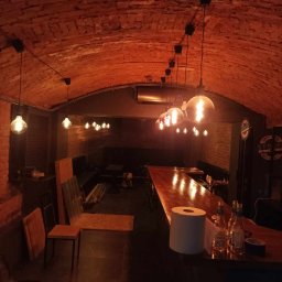 ROBERT MYKYTA RMD - Klimatyczne wnętrze lokalu z ceglanym sklepieniem łukowym, oświetlone wiszącymi żarówkami typu Edison, z drewnianym blatem baru w trakcie prac wykończeniowych, widoczne butelki i rolka papieru.