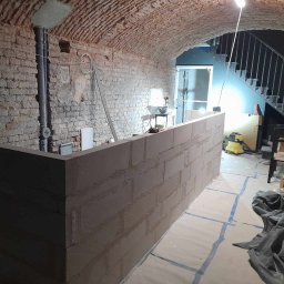 ROBERT MYKYTA RMD - Wnętrze piwnicy z ceglanym sklepieniem łukowym, w trakcie budowy baru z betonowych bloczków, widoczne narzędzia i materiały budowlane, zabezpieczona podłoga.