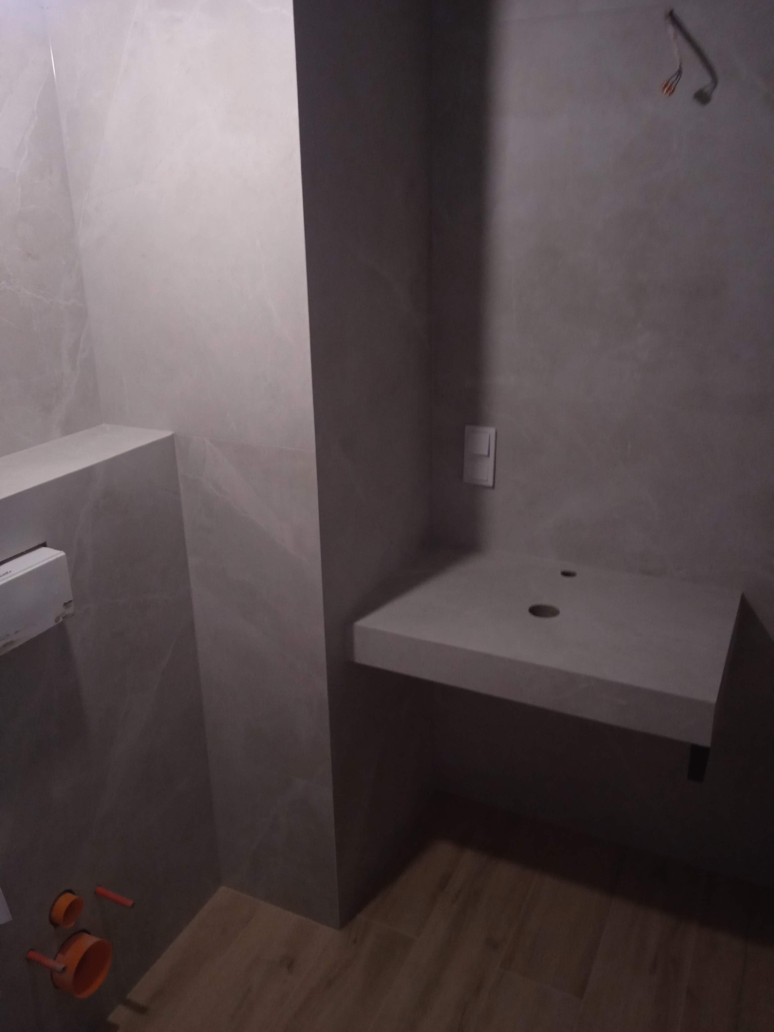 Łazienka w trakcie wykańczania z zamontowaną, kamienną umywalką podwieszaną. Widoczne przyłącza hydrauliczne, płytki imitujące kamień na ścianach i drewnopodobne panele na podłodze. Część kabli...