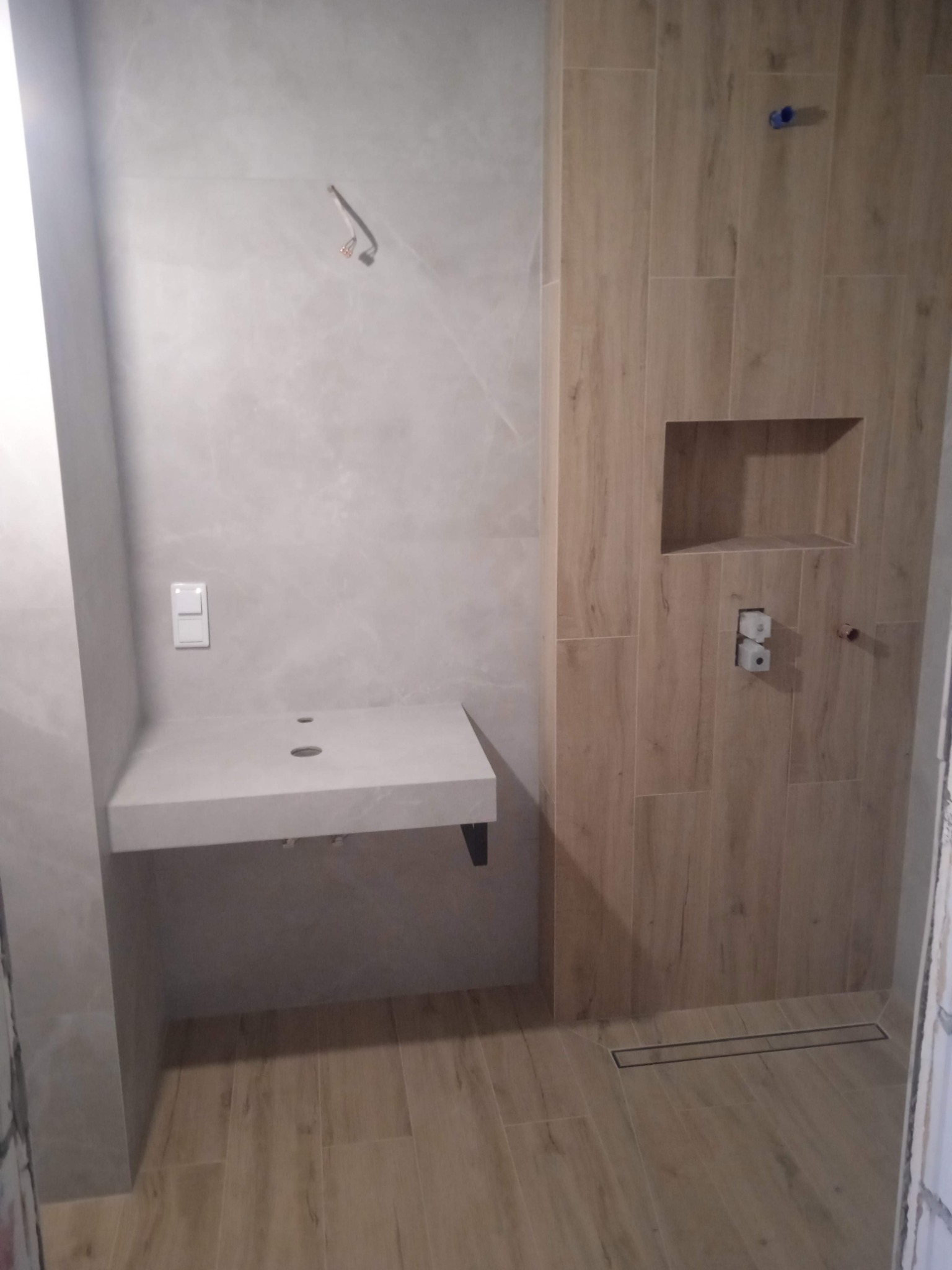 Nowoczesna łazienka po remoncie z drewnopodobnymi płytkami, wnęką prysznicową, odpływem liniowym i minimalistyczną umywalką z blatem z otworem na baterię.