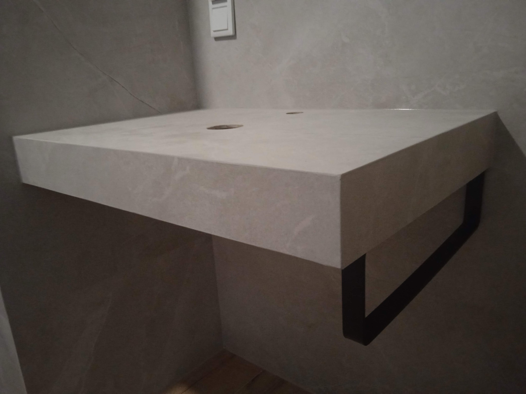 Półka umywalkowa z jasnego kamienia z dwoma otworami, wsparta na czarnym, metalowym stelażu w łazience o ścianach wykończonych płytkami imitującymi kamień.