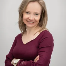 Portret uśmiechniętej kobiety z blond włosami, ubranej w bordową bluzkę z koronkowymi wstawkami na rękawach i zegarkiem na ręce, stojącej na jasnym tle.