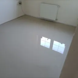 Świeżo wylana, samopoziomująca wylewka podłogowa w pustym pomieszczeniu z grzejnikiem i taśmą dylatacyjną przy ścianach, odbijająca okno.