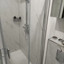 Nowoczesna kabina prysznicowa z białymi płytkami imitującymi marmur, chromowana armatura i czarny druciany koszyk na kosmetyki.