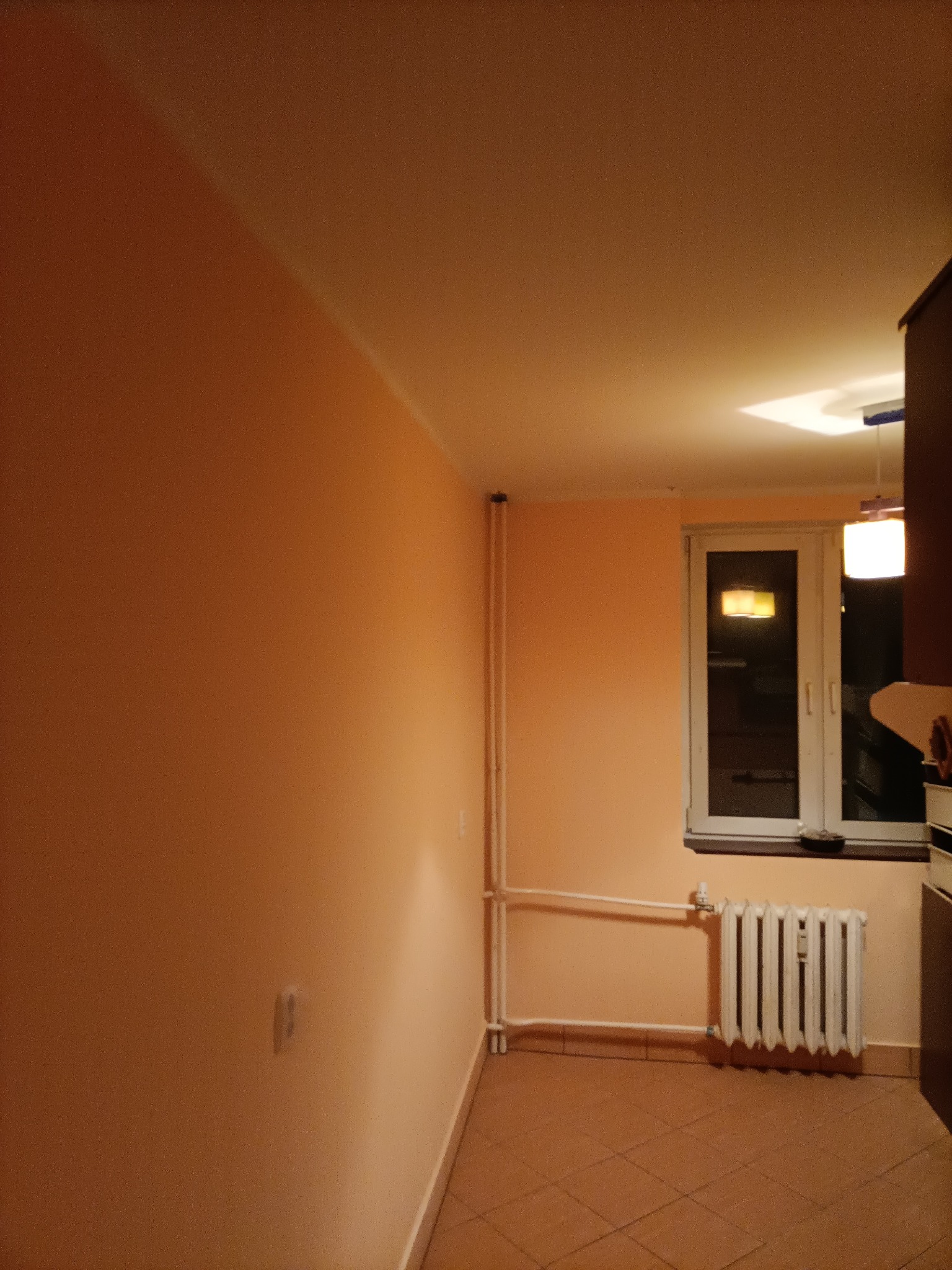 Pomalowane na brzoskwiniowo wnętrze pokoju z grzejnikiem pod oknem i płytkami na podłodze, widoczne rury grzewcze.