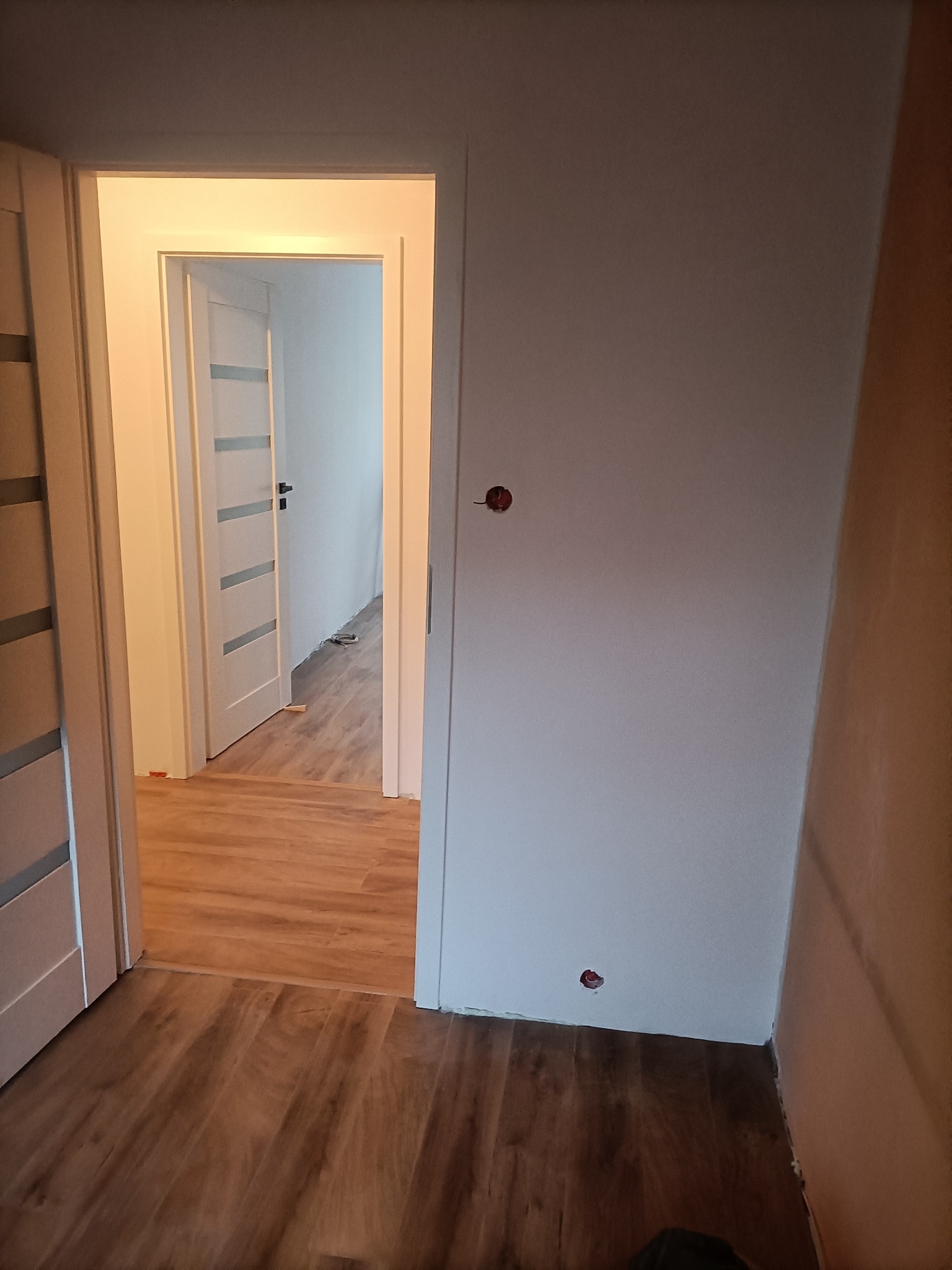 Wnętrze mieszkania w trakcie prac wykończeniowych: widok na korytarz z drewnopodobnymi panelami podłogowymi, białe ściany z widocznymi otworami po gniazdkach elektrycznych i częściowo zamontowane...