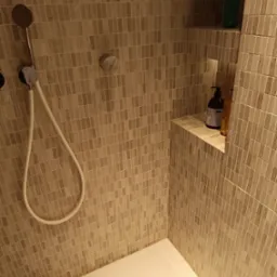 Wnętrze kabiny prysznicowej wyłożonej mozaiką w odcieniach beżu, z półkami wbudowanymi w ścianę, na których stoją butelki z kosmetykami i szamponami.