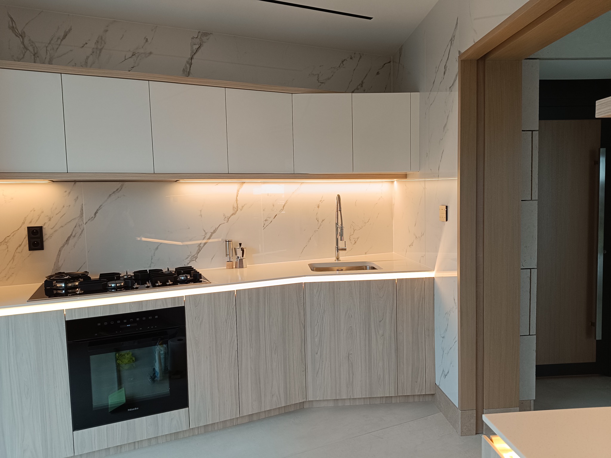 Nowoczesna kuchnia z białymi szafkami, marmurowym blatem i oświetleniem LED pod szafkami, z widoczną płytą grzewczą i piekarnikiem Miele.