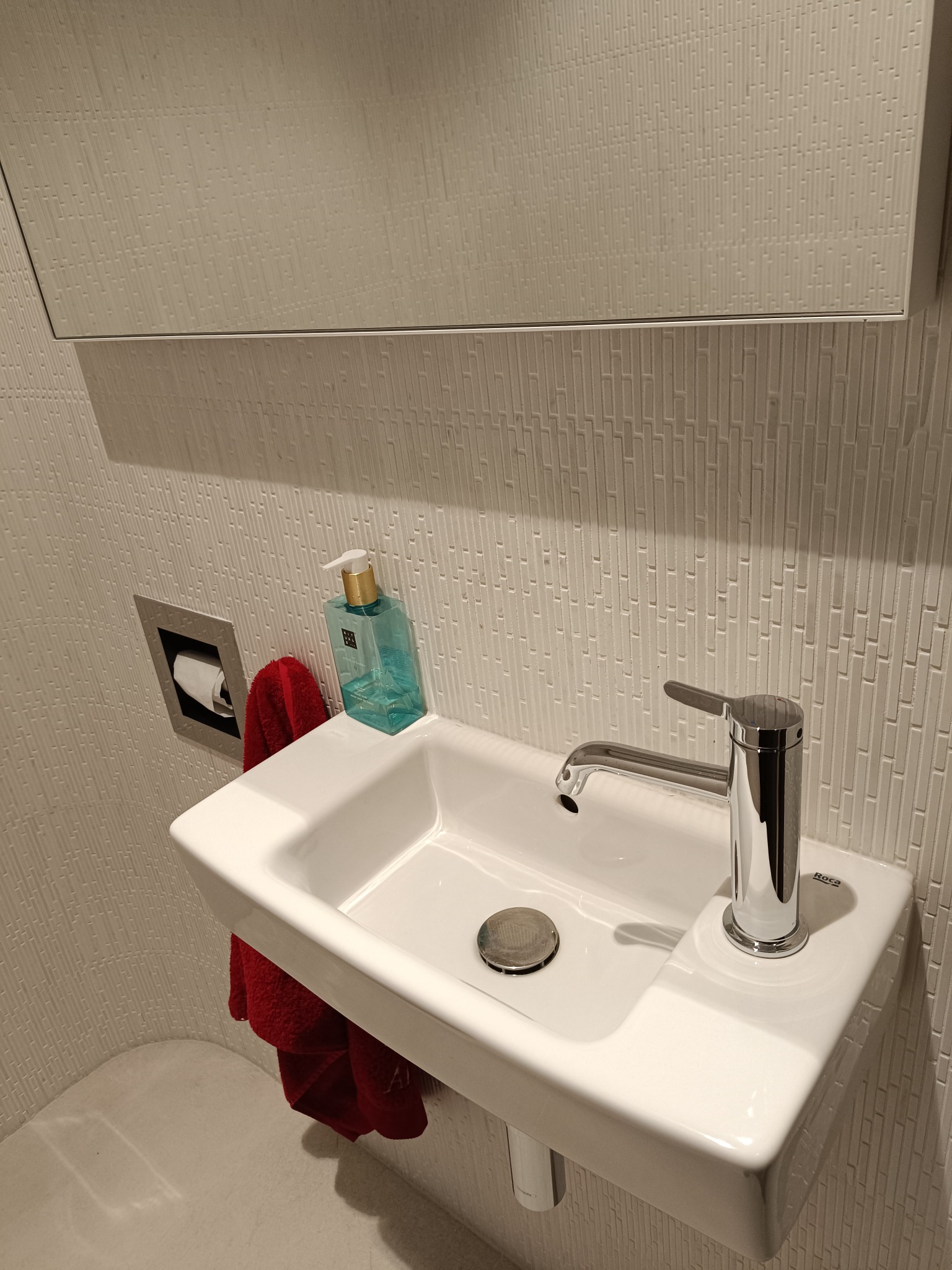 Mała, biała umywalka Roca z chromowaną baterią, dozownikiem mydła i czerwonym ręcznikiem na tle ściany z teksturą cegiełek.