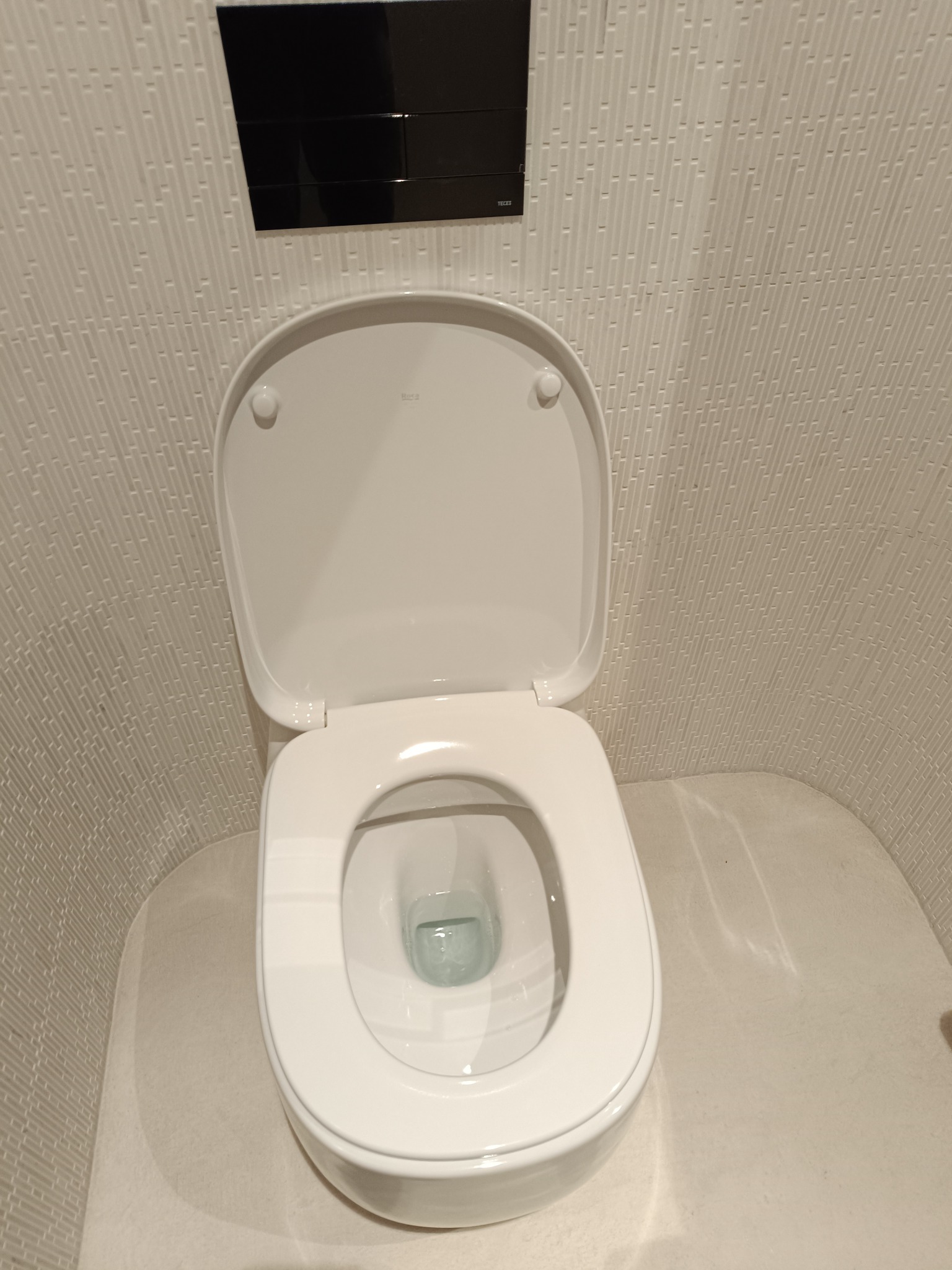 Biała toaleta z otwartą klapą, zamontowana na ścianie z dekoracyjnymi, pionowymi płytkami i czarnym panelem spłukującym.