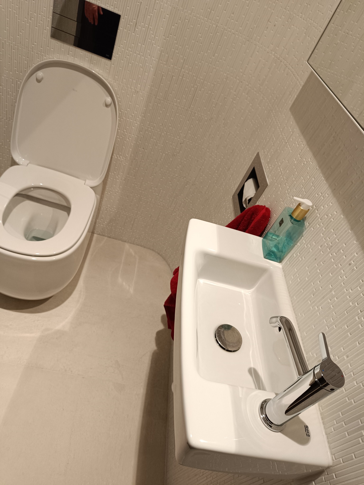 Mała, nowoczesna łazienka z białą ceramiką: wisząca toaleta z otwartą klapą, prostokątna umywalka z chromowaną armaturą Roca, czerwony ręcznik i dozownik mydła na tle ściany wyłożonej teksturą...