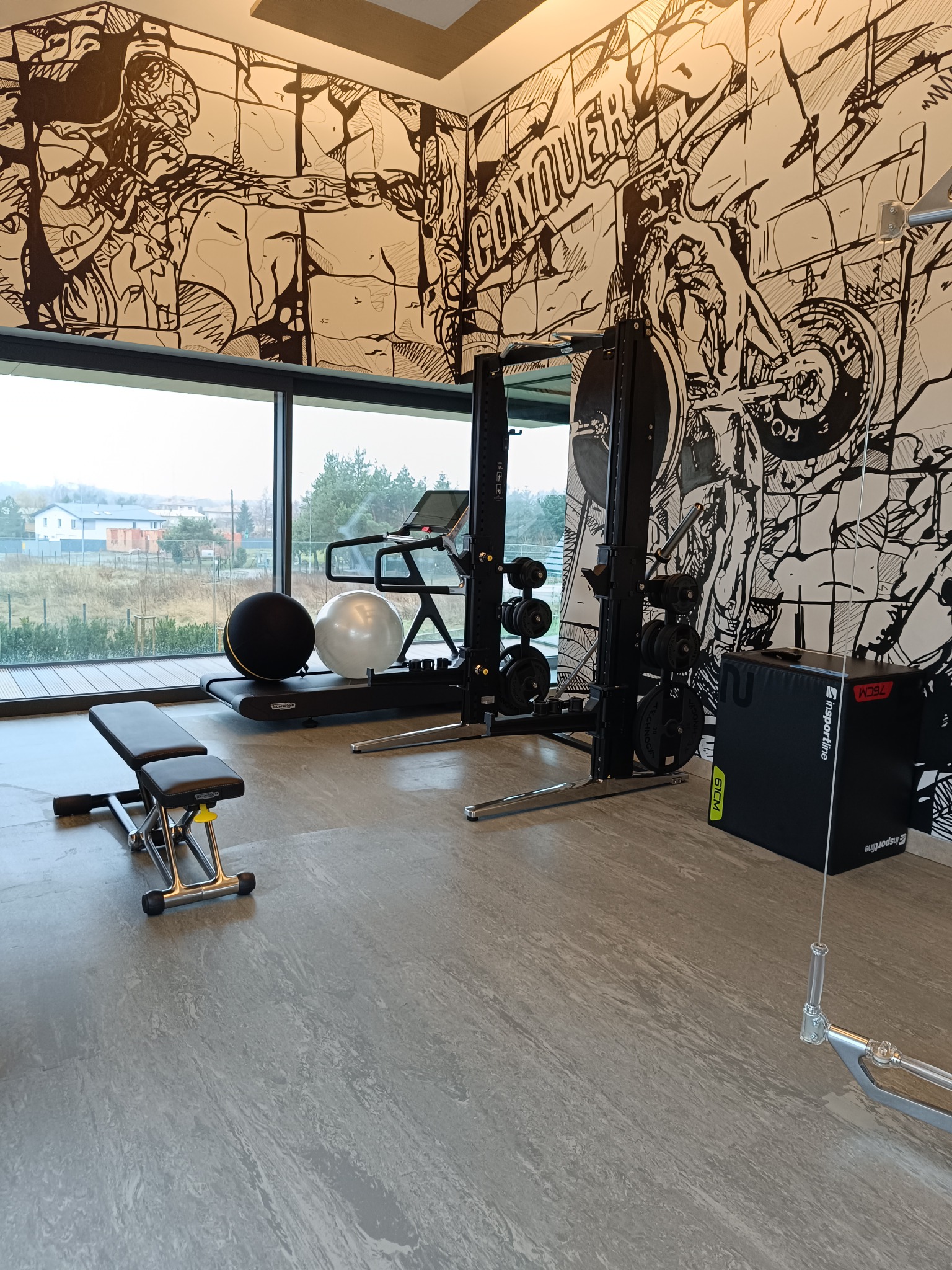 Nowoczesna siłownia domowa z bieżnią, atlasem do ćwiczeń, ławką i piłkami gimnastycznymi na tle ściany pokrytej czarno-białym muralem o tematyce sportowej.