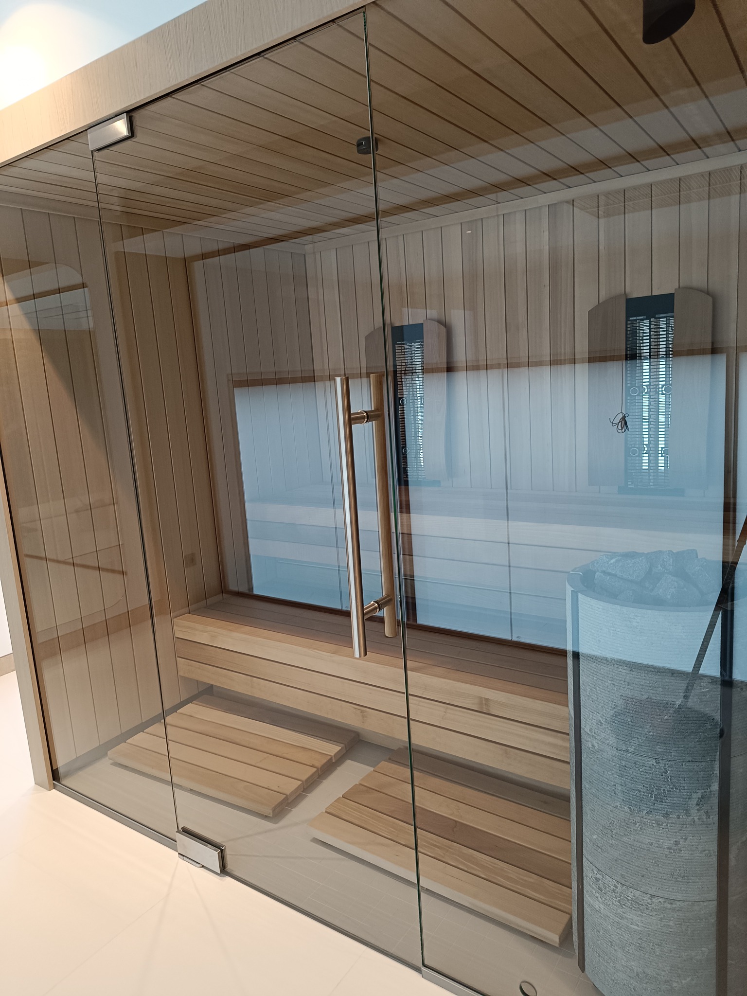 Szklana sauna z drewnianym wnętrzem, widoczne ławki, kamienie w kamiennym pojemniku i grzejniki na ścianie.