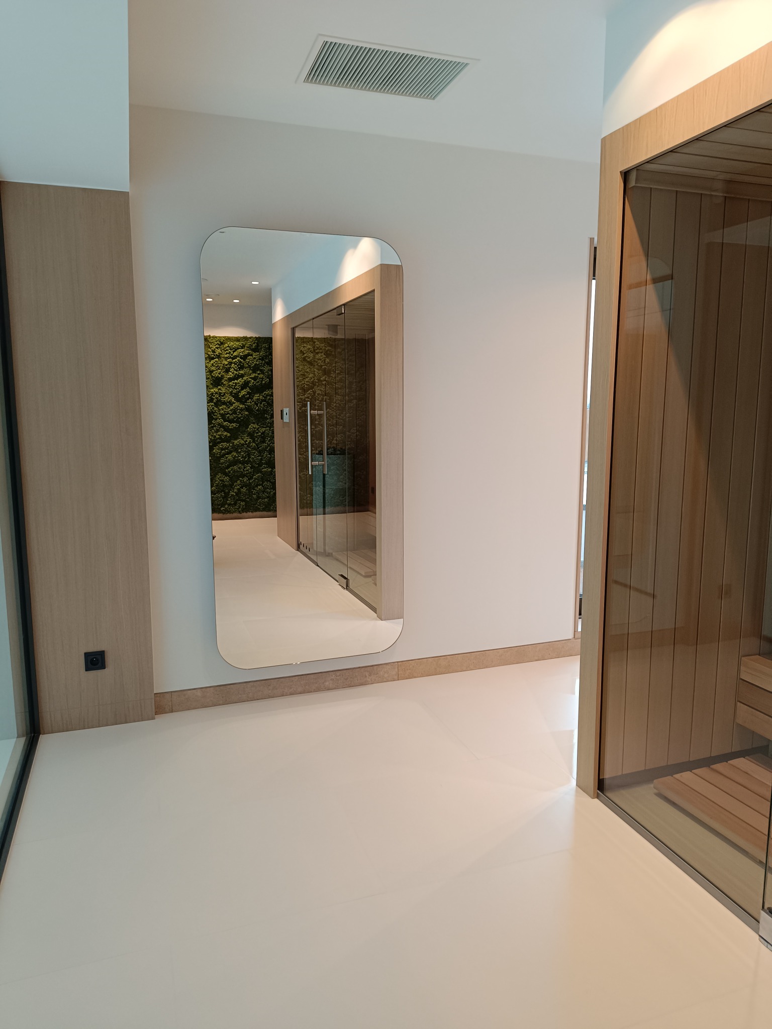 Nowoczesne wnętrze z sauną i ścianą z mchu, widoczne w lustrze o zaokrąglonych krawędziach, biała podłoga i drewniane wykończenia.