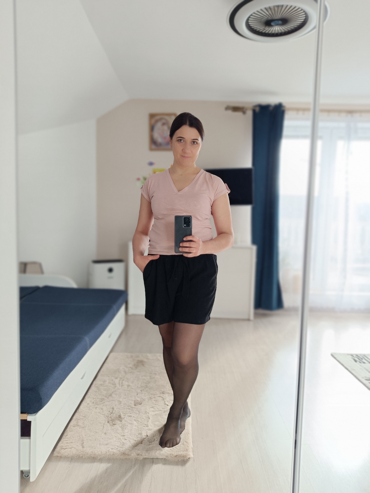 Osoba robiąca selfie w lustrze w schludnym pokoju z łóżkiem z szufladami, dywanem i widokiem na okno z zasłonami.