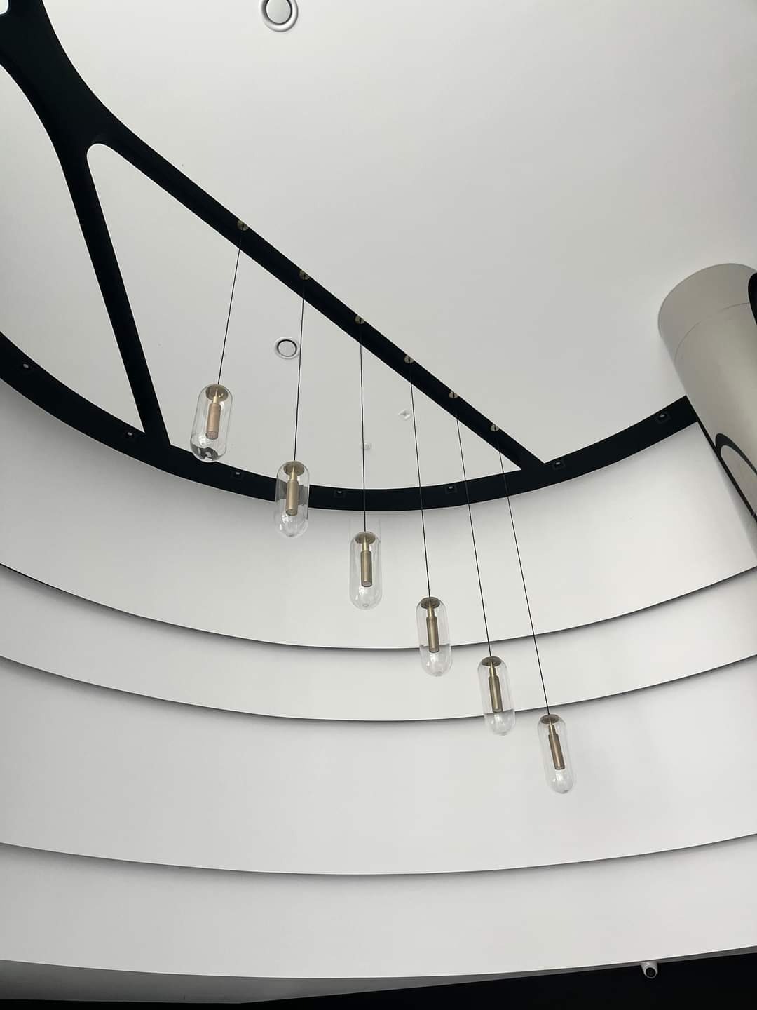 Nowoczesny sufit z kaskadowymi, szklanymi lampami w kształcie kapsuł, podwieszonymi na cienkich linkach, z widoczną dekoracyjną czarną konstrukcją i zaokrąglonym elementem architektonicznym.