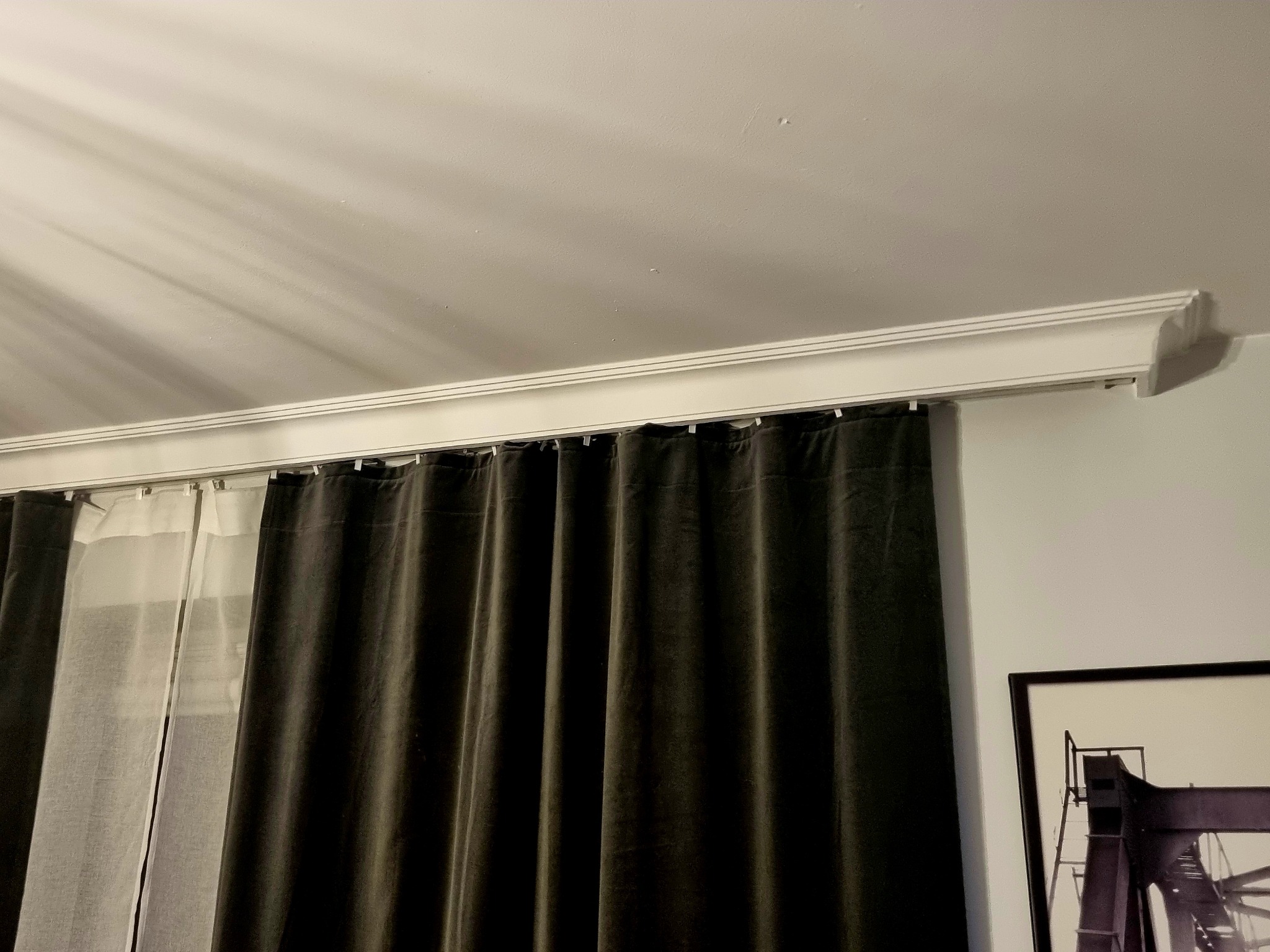 Fragment wnętrza z ciemnymi zasłonami wiszącymi na karniszu z dekoracyjną listwą przysufitową, obok rama obrazu z motywem industrialnym.