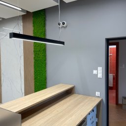 Electro4you - Nowoczesne biuro z recepcją: długa, jasna lada, oświetlenie LED, ściana z mchu i marmuru. Dwie kamery bezpieczeństwa nad ladą. Widoczne włączniki światła.